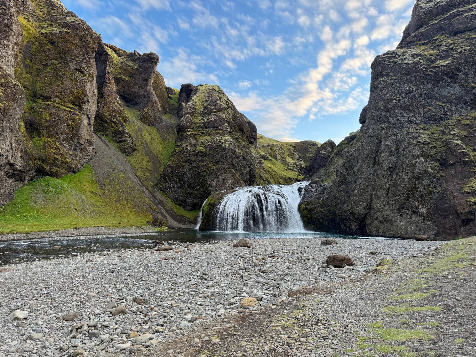 A small waterfall between mossy rock walls, with a gravel riverbed in the foreground