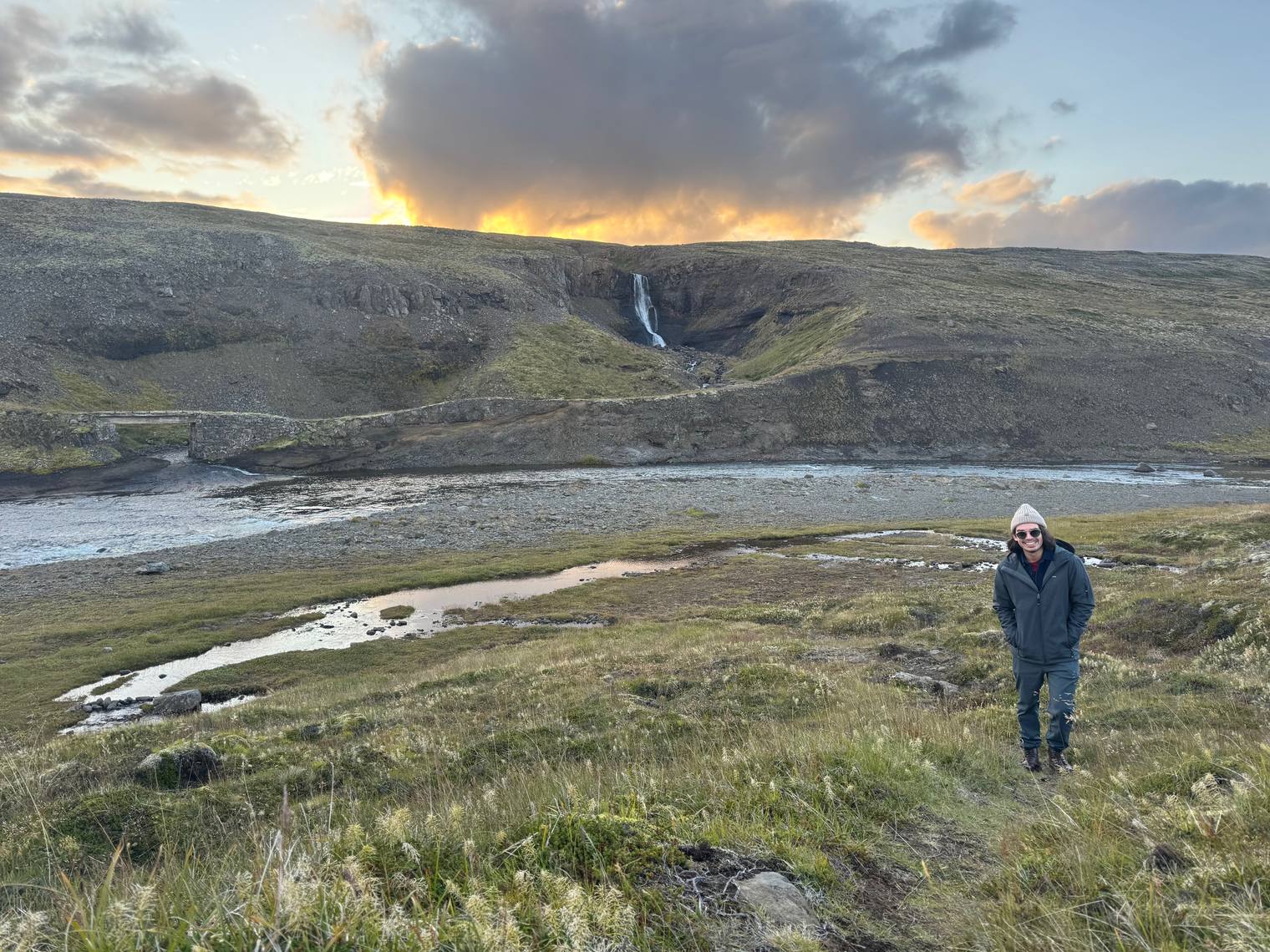 A distant waterfall on a highland hillside at sunset, with a person standing in the grass