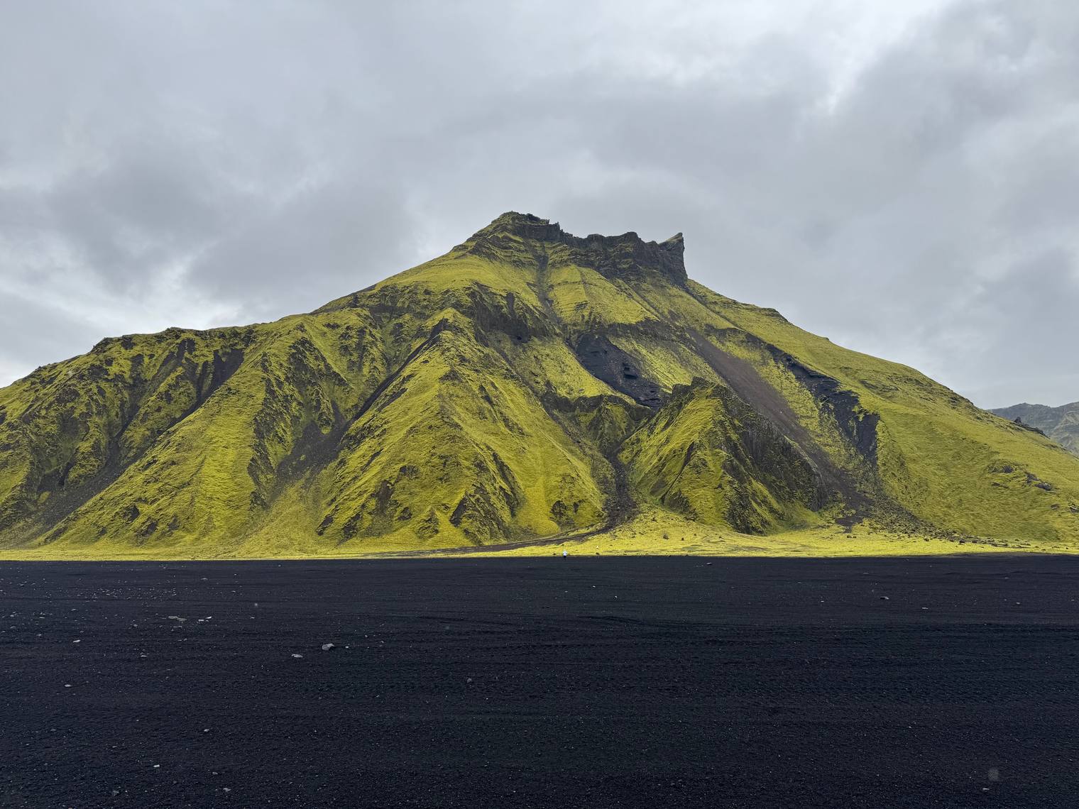 A moss-covered mountain rising above a plain of black sand under an overcast sky