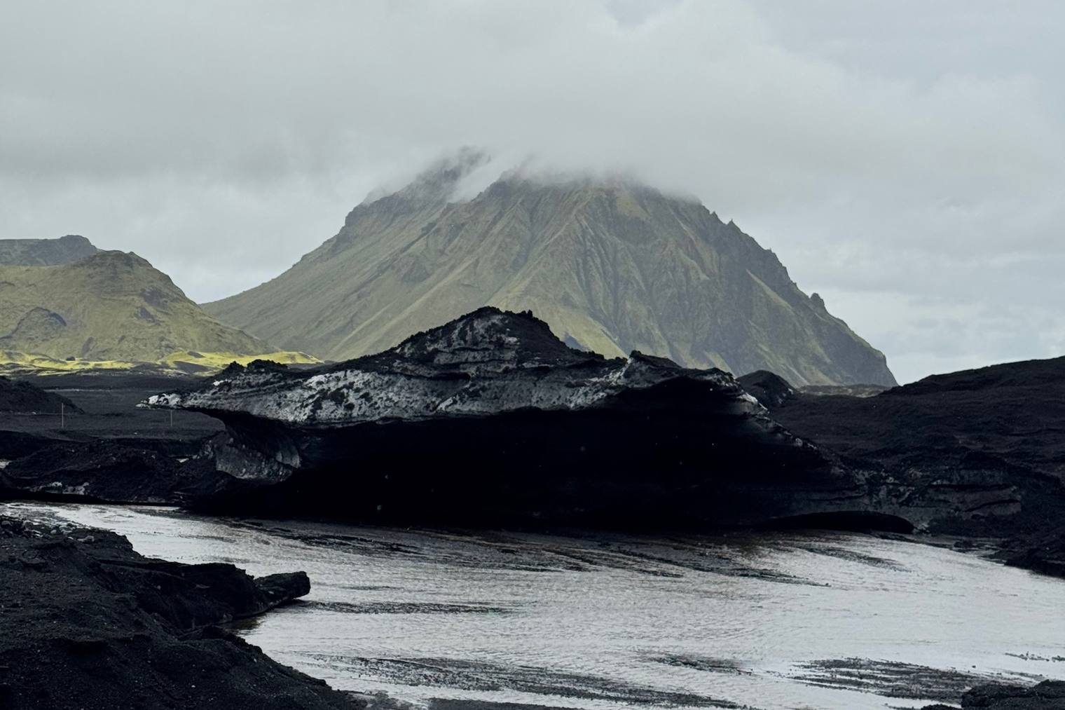 A cloud-capped mountain above a black glacial river bed